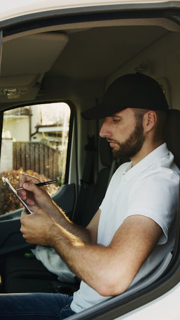 A man wearing a cap, dark jacket, and trousers is engaged in packing or organizing cardboard boxes labeled 'FRAGILE' outside a building for a home relocation process. He stands next to a large stack o